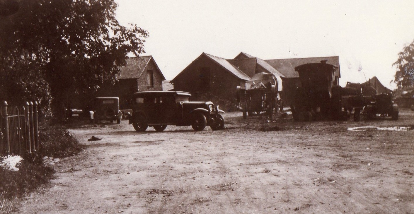 An image of church farmyard from times gone by. A threshing machine is running and early cars are parked nearby